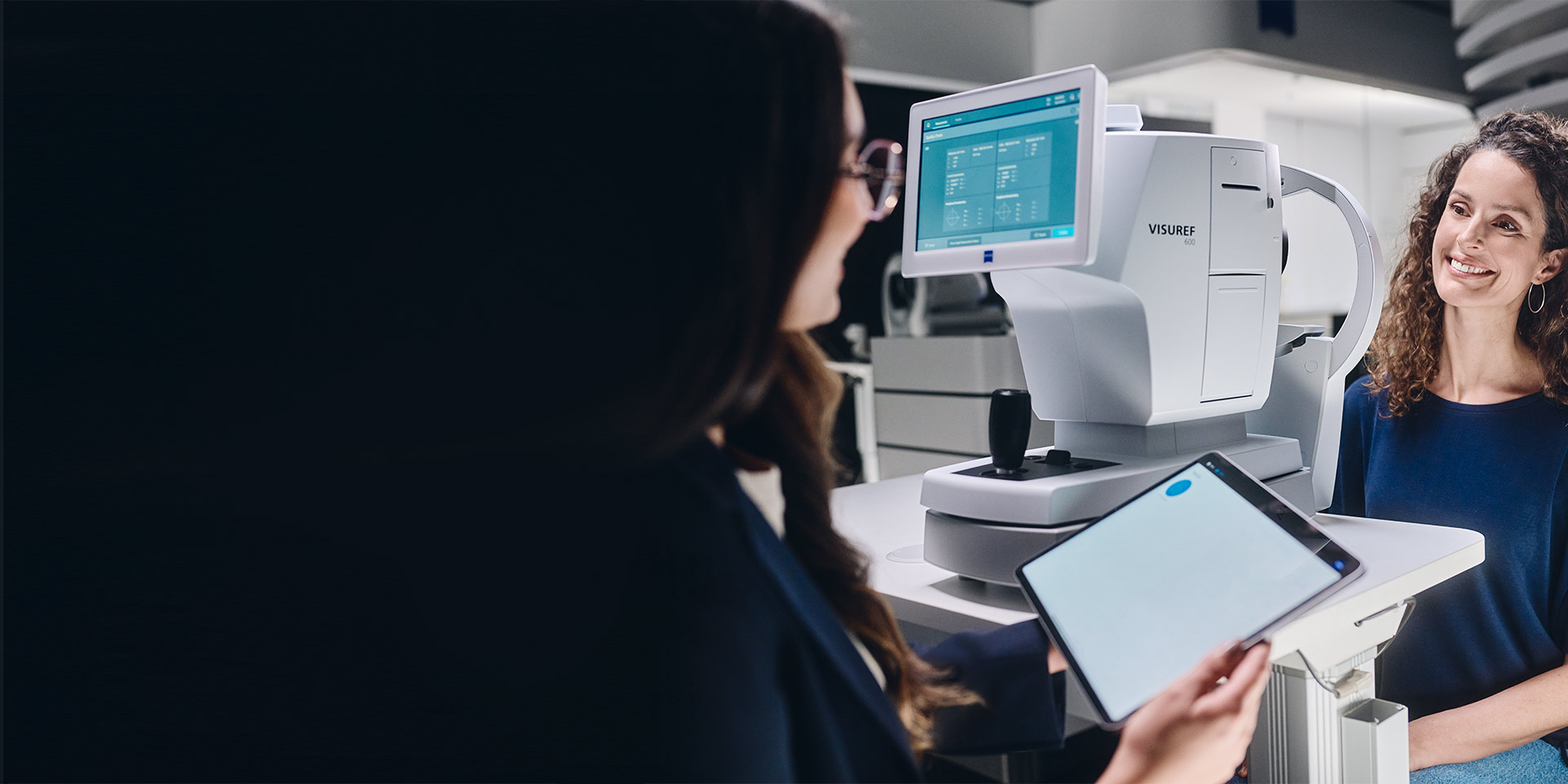 A female eye care professional looking at her tablet sitting in front of the ZEISS VISUREF 600 and talking to her customer.