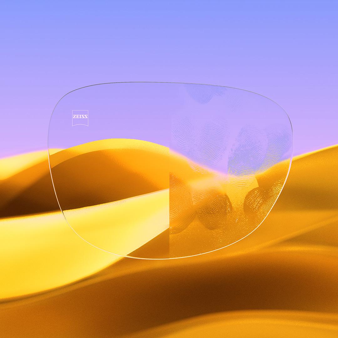 A split eyeglass lens with one side being clear and clean, featuring ZEISS DuraVision Gold UV and one side with fingerprints covering the lens.