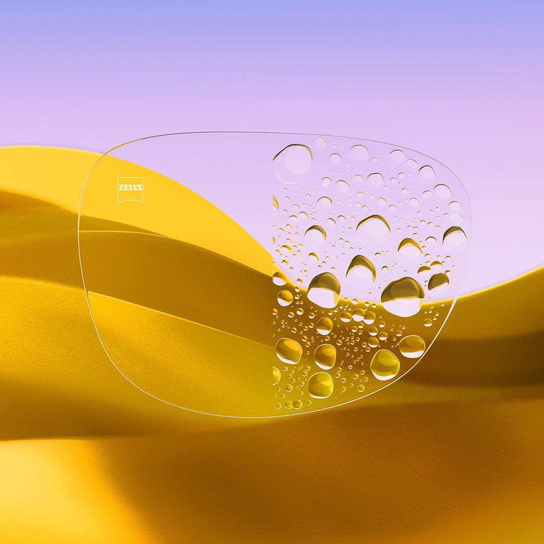 A split eyeglass lens with one side being clear and clean, featuring ZEISS DuraVision Gold UV and one side with water drops covering the lens.