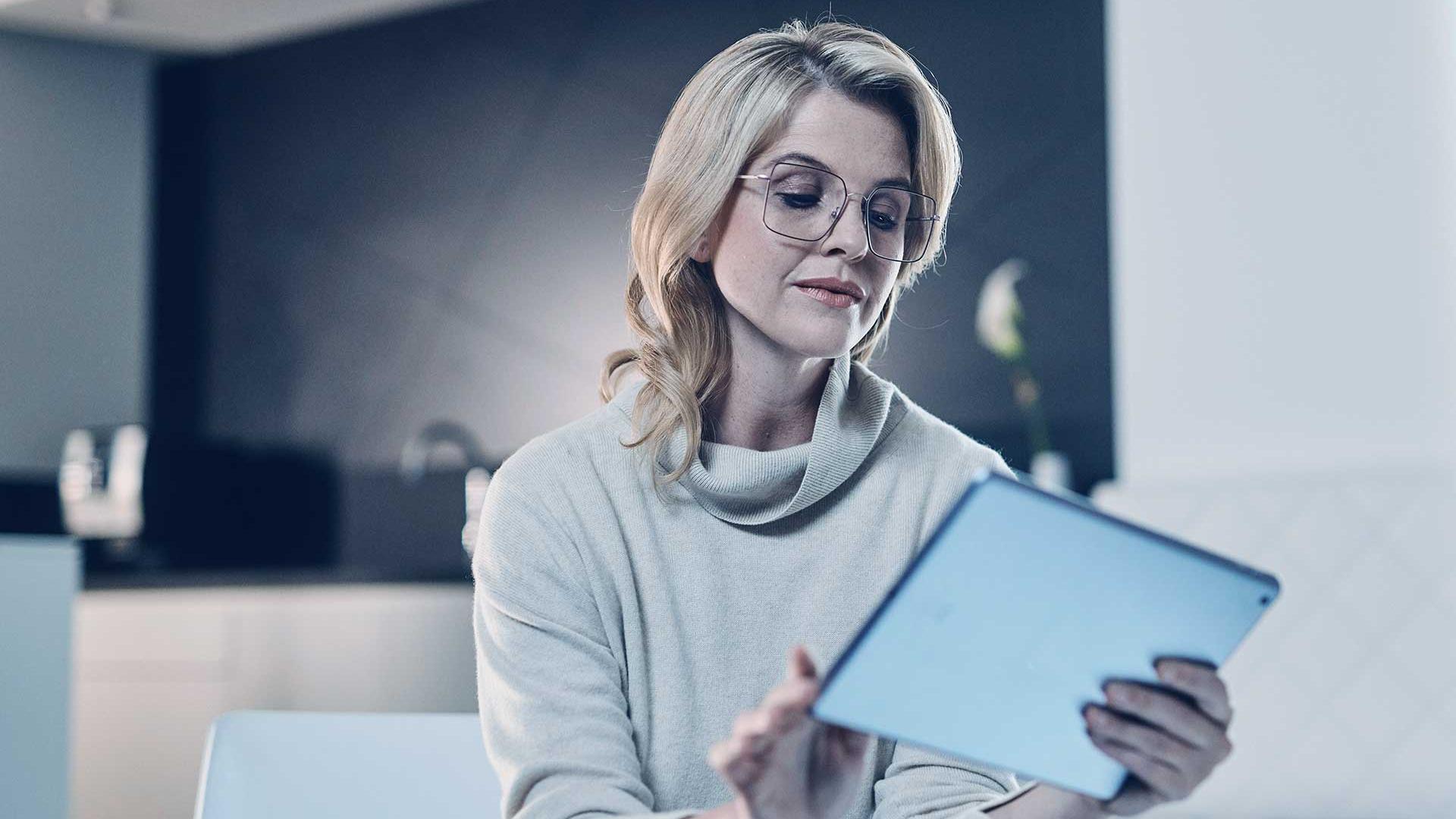 Femme blonde portant des lunettes et un pull beige tient une tablette en main et lit