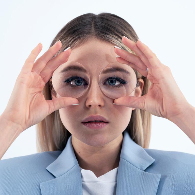 Une jeune femme blonde tient des verres devant ses yeux pour montrer l’effet « fisheye » causé par les verres épais.