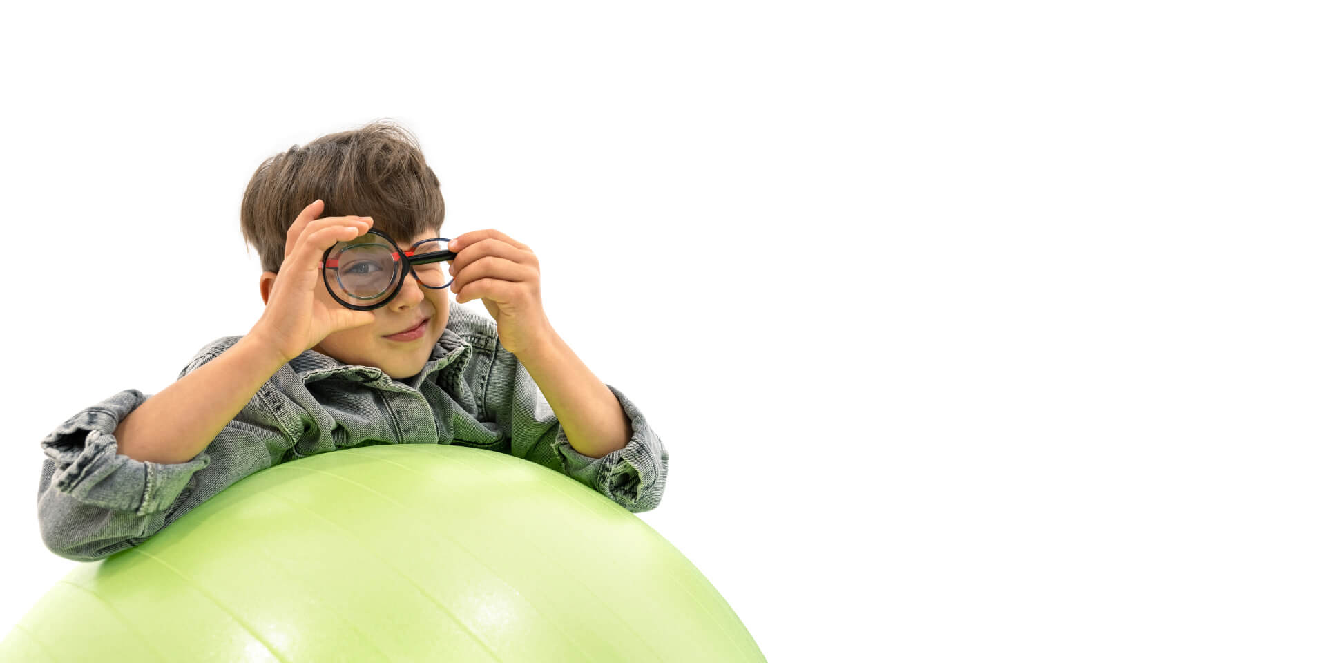 Un garçon porte des verres ZEISS Myopia Management, appuyé contre un ballon de gymnastique et tenant une loupe devant un œil.