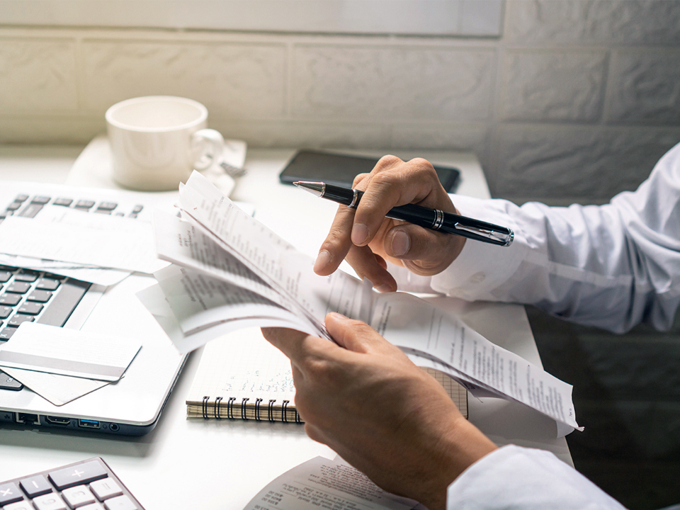 Scène de bureau typique, avec une personne occupée à examiner des feuilles de calcul et des documents. Seuls les bras de la personne sont visibles ; elle porte dans les mains une pile de feuilles de calcul. On voit en arrière-plan un bureau sur lequel se trouve un ordinateur portable. Une fenêtre en haut à gauche de l’écran montre un homme. Il a les cheveux et la barbe grise, il porte des verres ZEISS OfficeLens et son expression faciale est neutre.