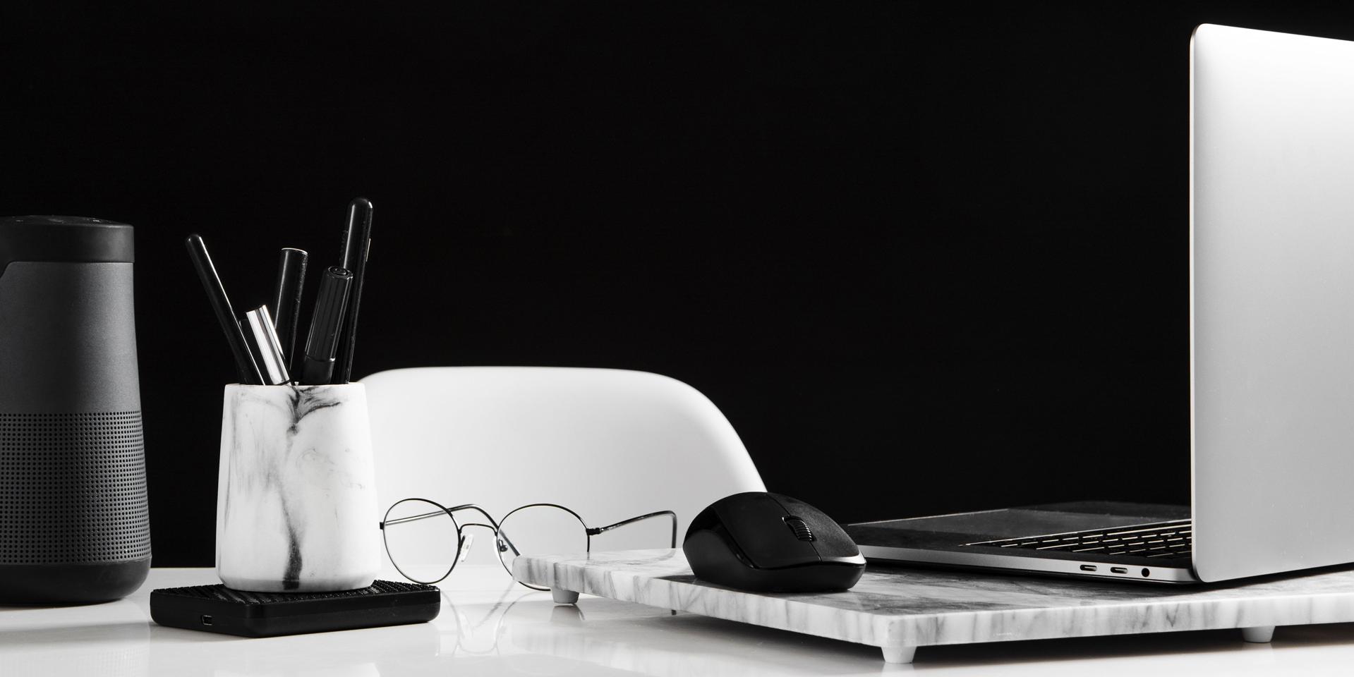 Tidy workspace showing an open laptop on the right side with a mouse. In front of it lies a pair of glasses. On the left side of the picture is a box with pens.