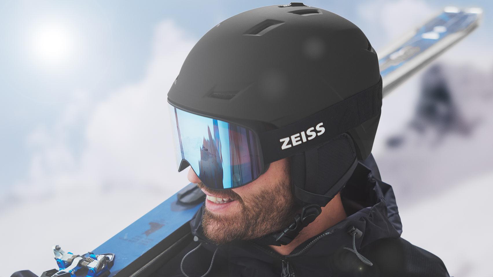 A man wearing a black helmet with blue mirrored ZEISS ski goggles, with skis over his right shoulder.