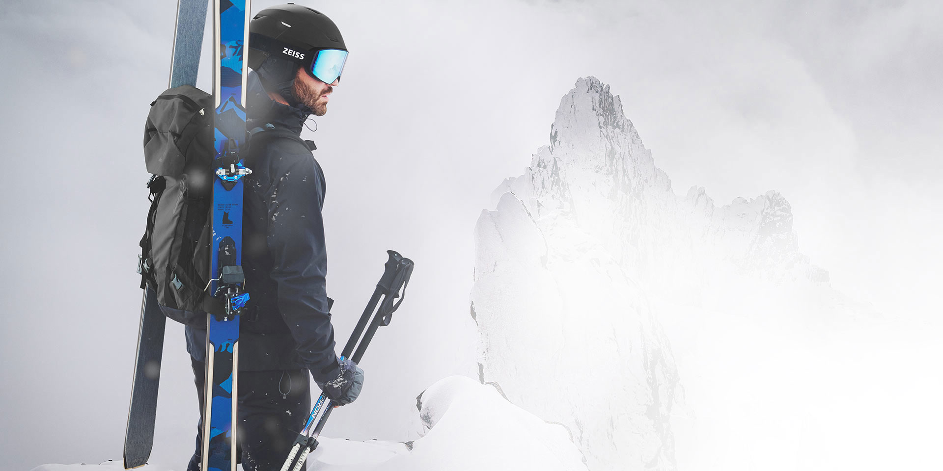 A man wearing a black helmet with blue mirrored ZEISS ski goggles. He has skis and a backpack on his back and is holding the ski poles in his right hand. He is standing on a mountain top.