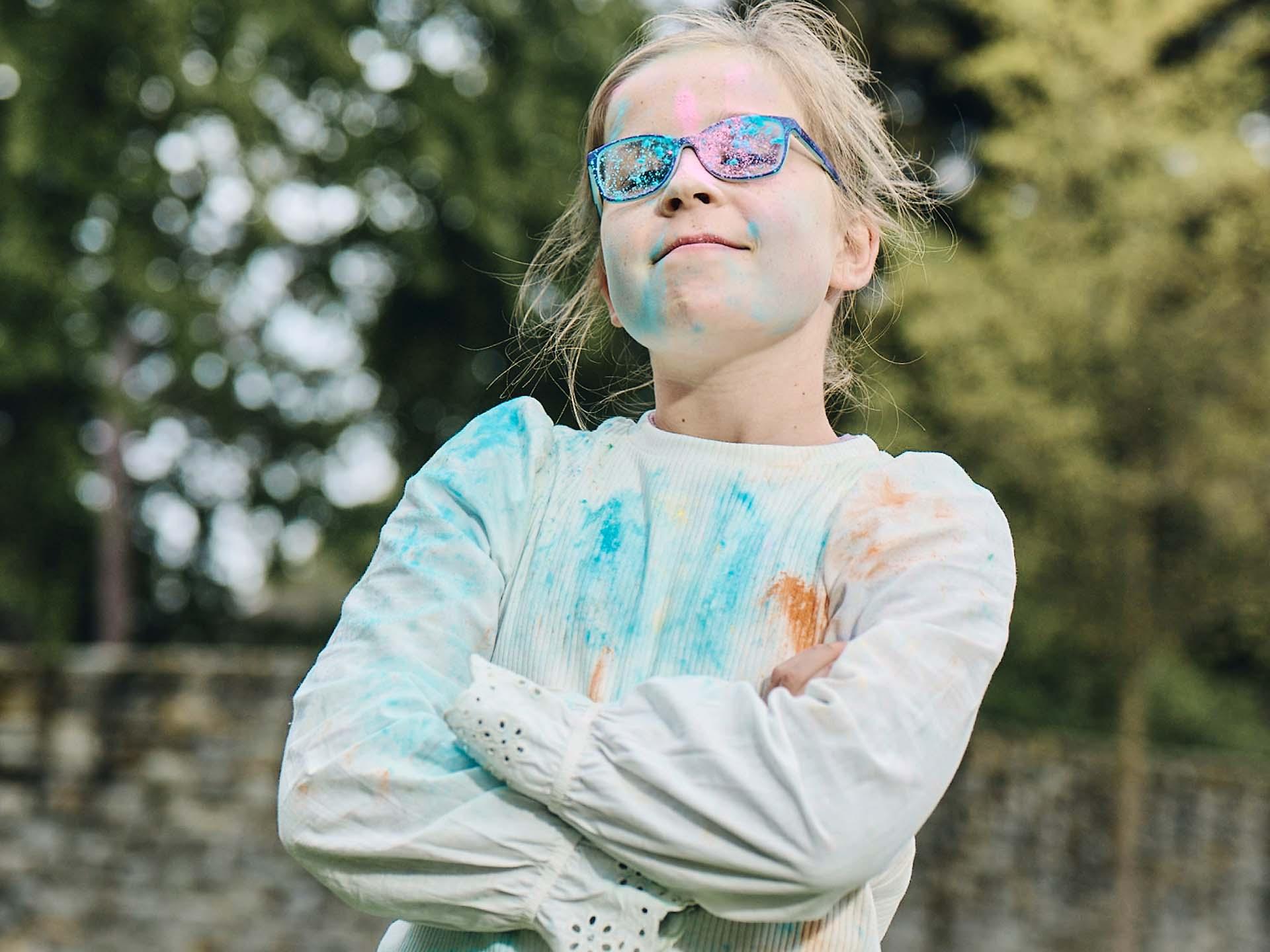 A girl with crossed arms and dirty glasses from playing with colored powder looks defiant and grins.
